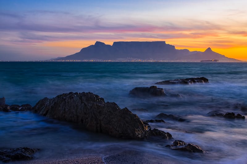 Der Tafelberg als Silhouette über der Bucht, vorne dunkle Felsen im Wasser, Himmel in Orange