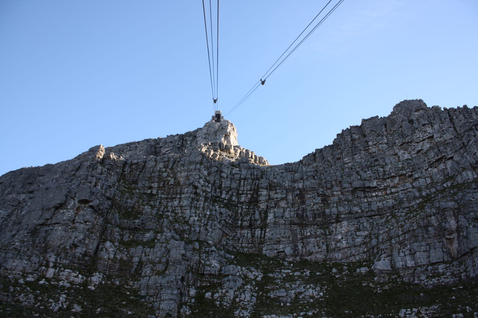 Zwei Tragseile der Seilbahn führen von unten über die graue Felswand zur Bergstation auf der Kante